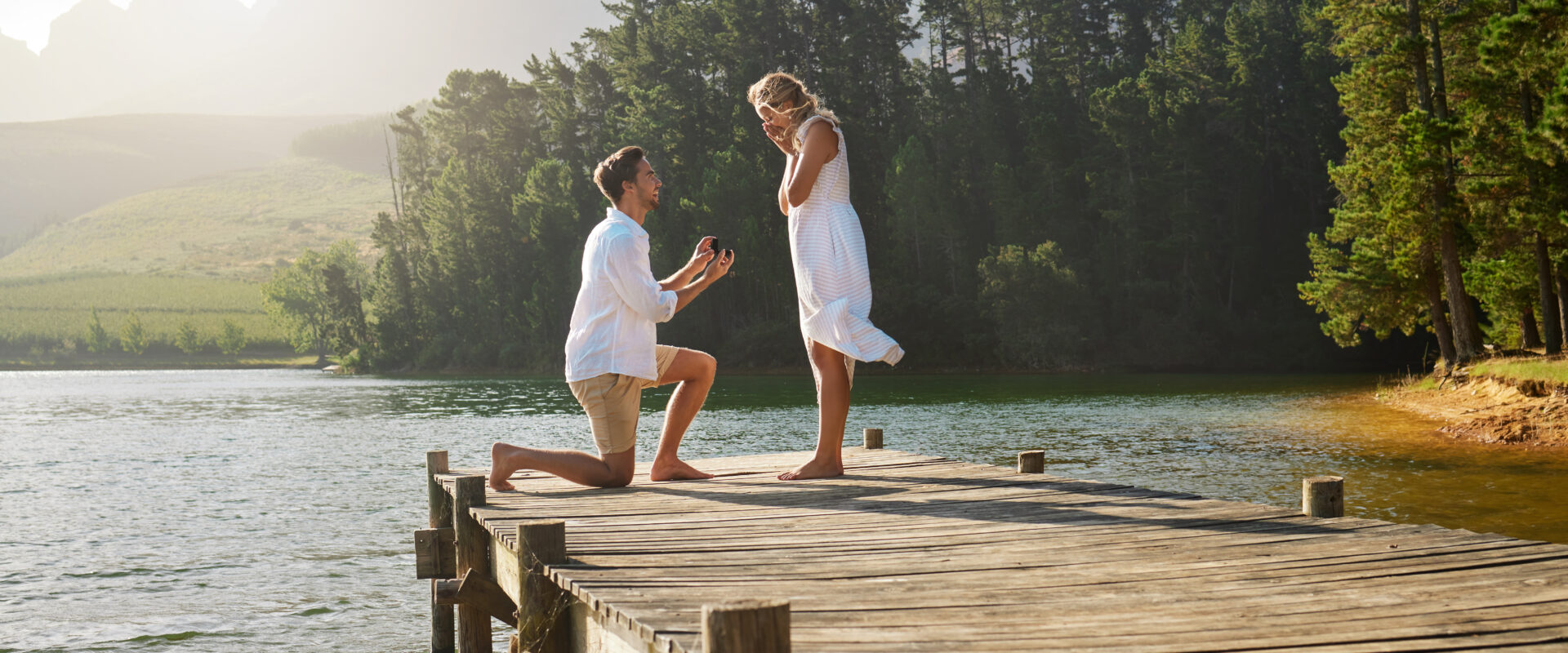 Man, woman and engagement proposal by lake on vacation with surprise, wow or happiness in sunshine. Couple, marriage offer and ring in nature for romance, love and happy on holiday in summer by water