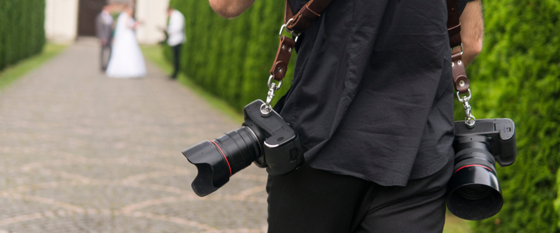 Professional wedding photographer takes pictures of the bride and groom in garden, the photographer in action with two cameras on a shoulder straps.