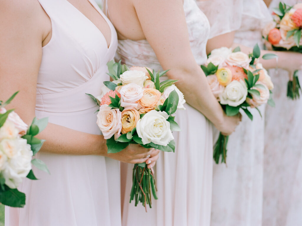 Bridesmaids hold colorful floral bouquets during an outdoor wedding ceremony in a soft, natural setting on a sunny day