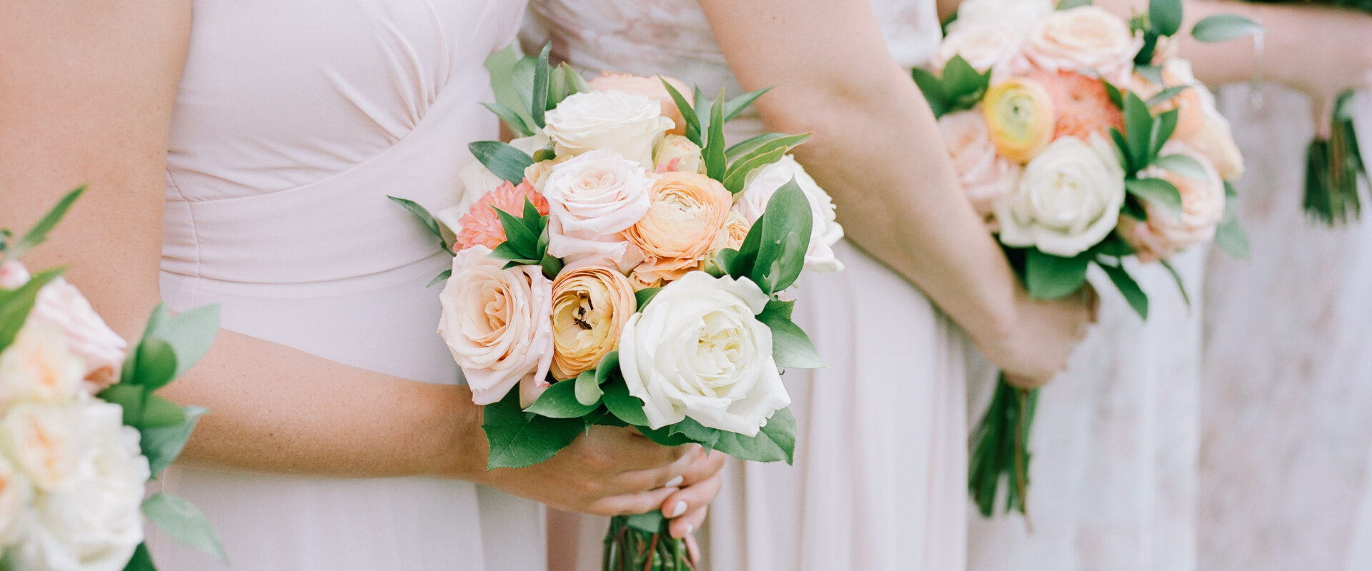 Bridesmaids hold colorful floral bouquets during an outdoor wedding ceremony in a soft, natural setting on a sunny day