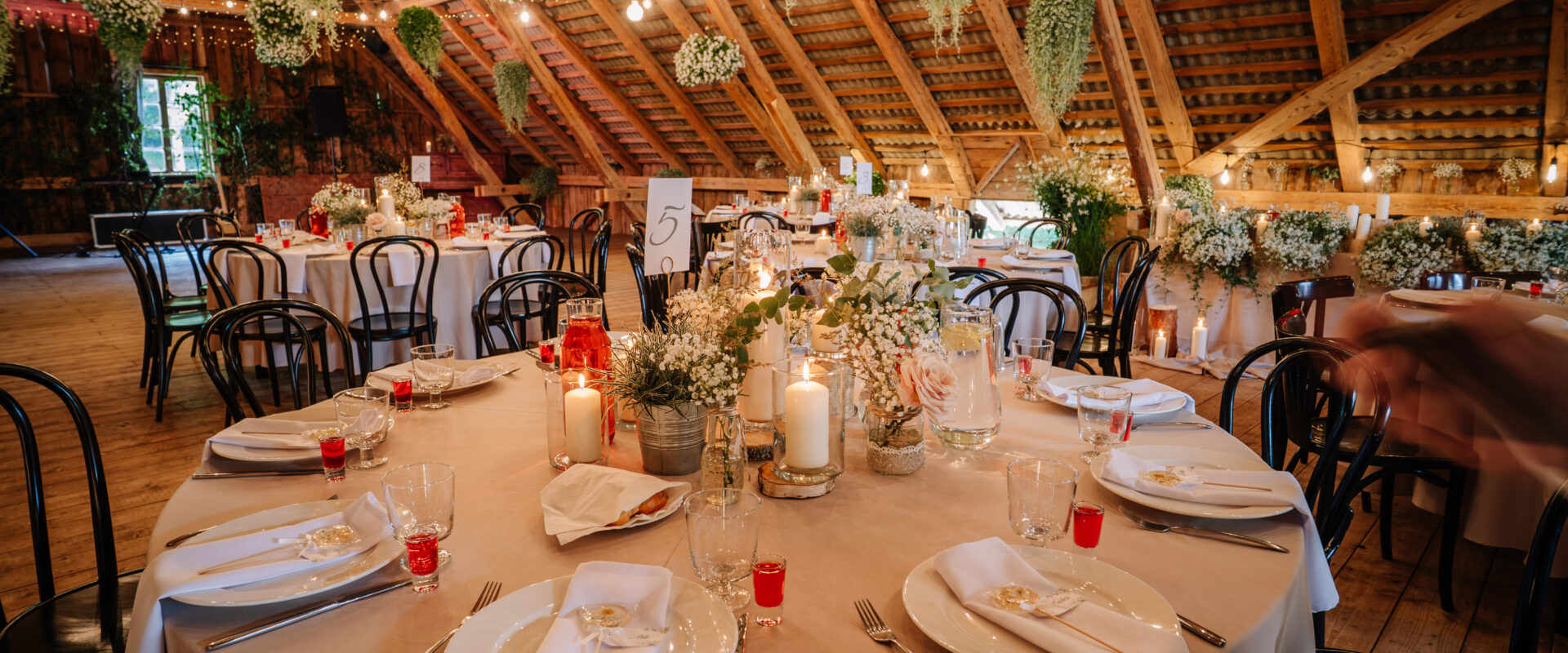 Rustic wedding reception setup with round tables, floral centerpieces, candles, string lights, hanging greenery, and elegant table settings in a wooden hall.