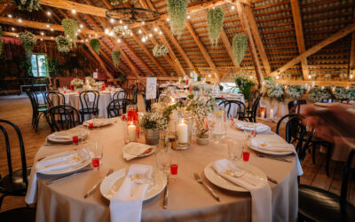 Rustic wedding reception setup with round tables, floral centerpieces, candles, string lights, hanging greenery, and elegant table settings in a wooden hall.